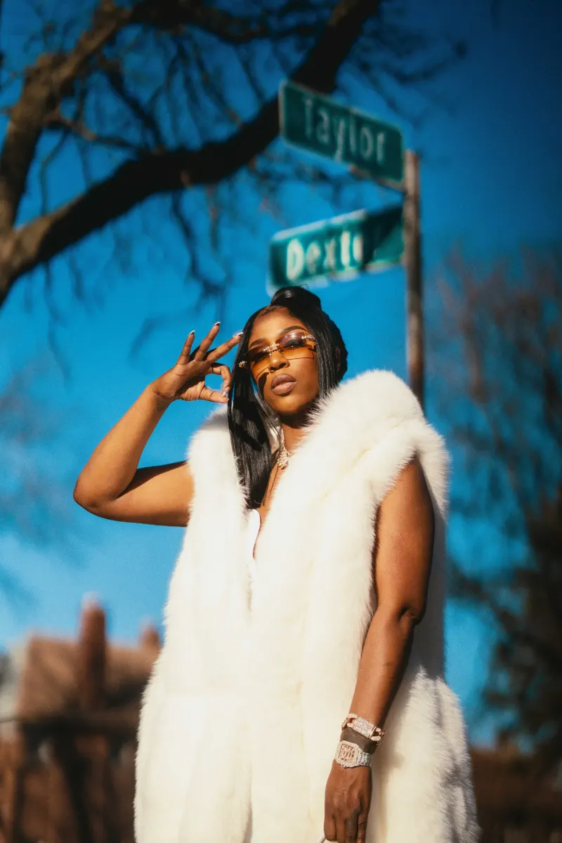 A woman in a white fur coat and sunglasses under a street sign, shot from below against a blue sky