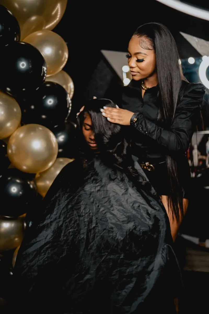 Hairstylist Kaniya J working on a client's long hair beside black and gold balloons