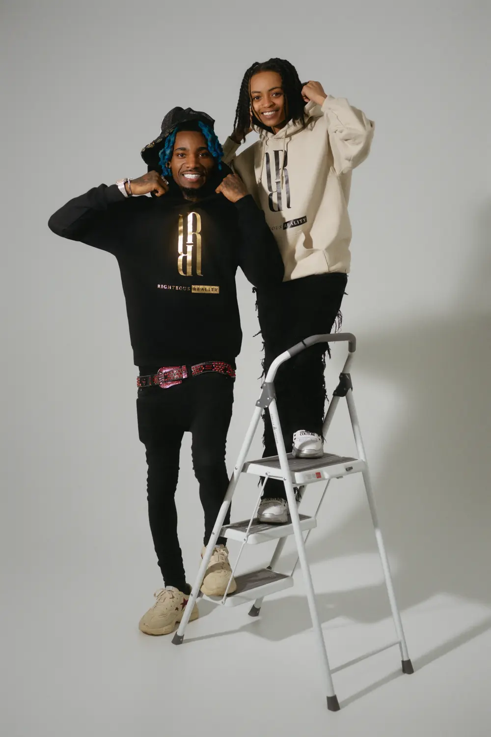 Two models in Righteous Reality hoodies, one black and one cream, posing on a silver step ladder