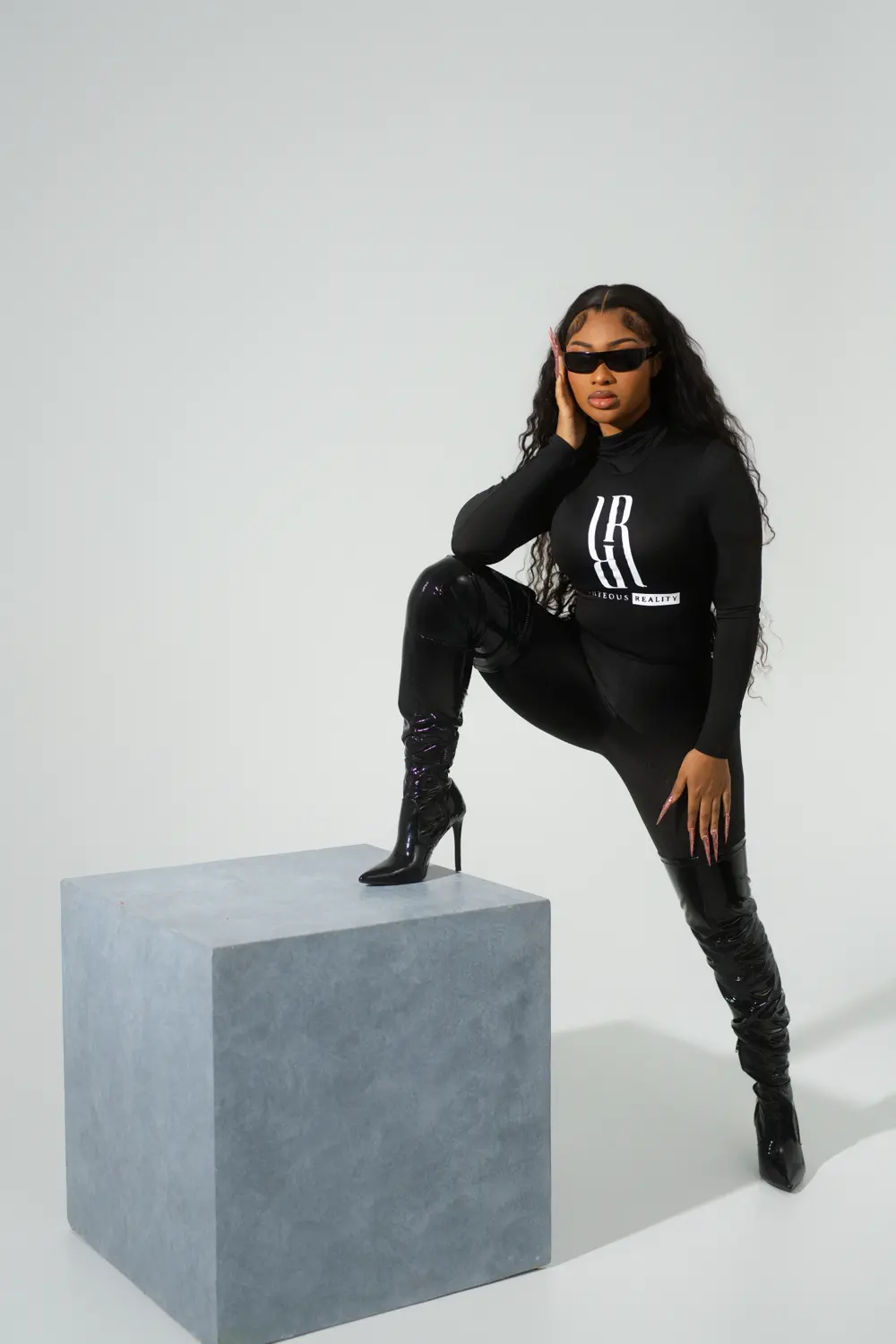 A model in a black Righteous Reality bodysuit with one heel up on a concrete cube