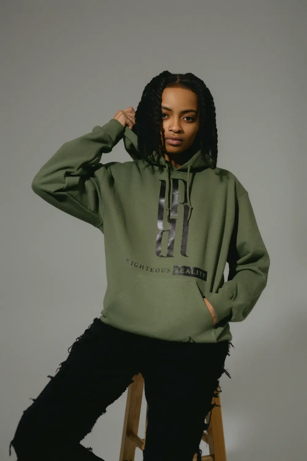 A model with braids in an olive Righteous Reality hoodie, one hand in the pocket, sitting on a wooden stool