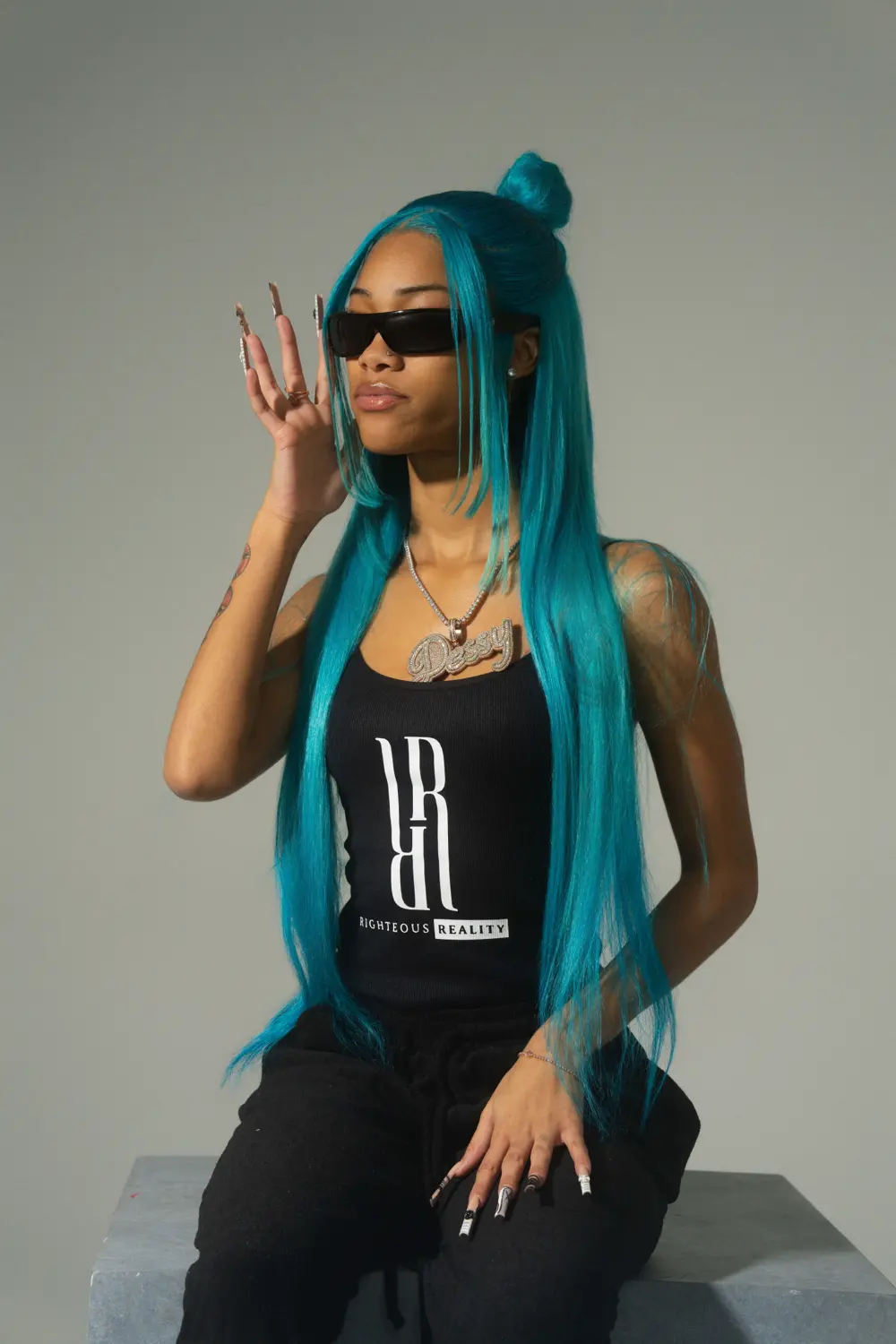 A model with turquoise hair in a black Righteous Reality tank top and sunglasses, sitting on a concrete block