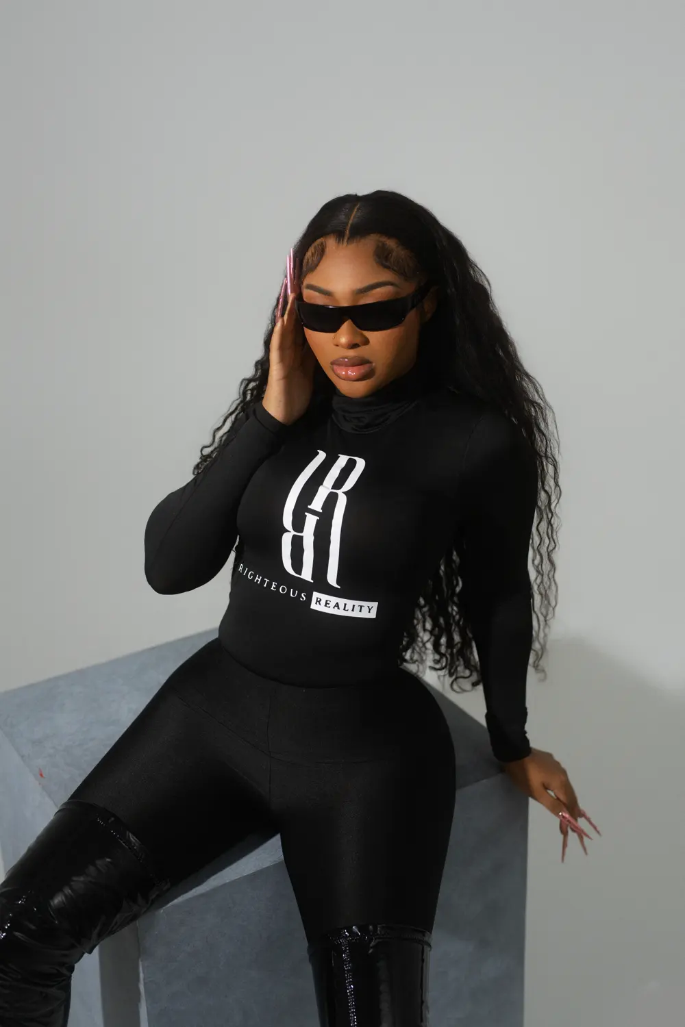 A model in a black Righteous Reality long sleeve leaning on a concrete cube, touching her sunglasses