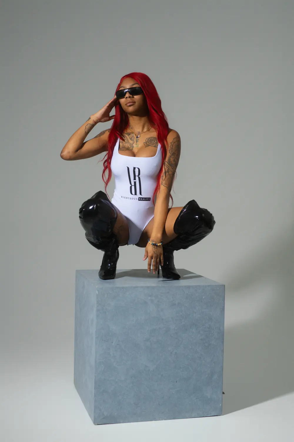 A model with red hair in a white Righteous Reality bodysuit, crouching on top of a concrete cube