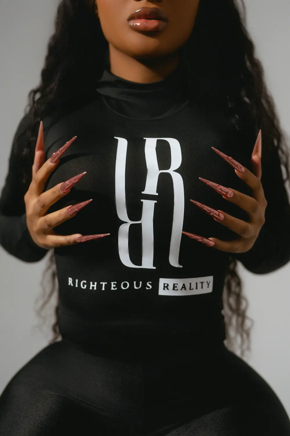 Close crop of the Righteous Reality logo on a black long sleeve, framed by long pink nails